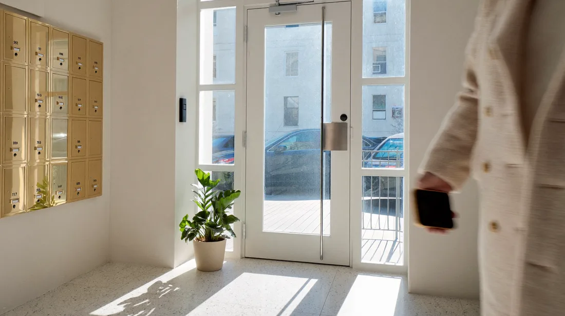 A sunlit residential lobby: a resident passing the letterboxes, phone in hand.